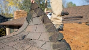 Roof replacement in progress as a worker removes worn shingles, revealing the underlying structure. Learn more about Roof Replacement Cost. 